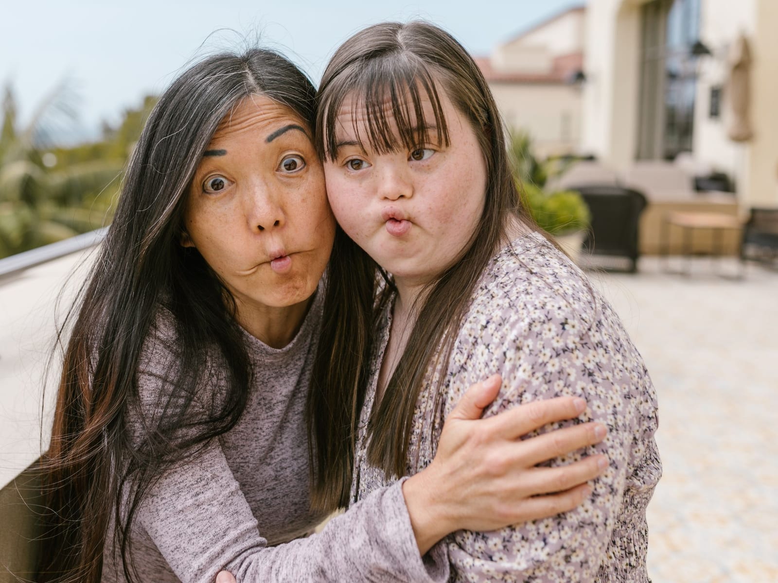 Two women lean their heads together and pull playful faces on a terrace surrounded by palm trees.