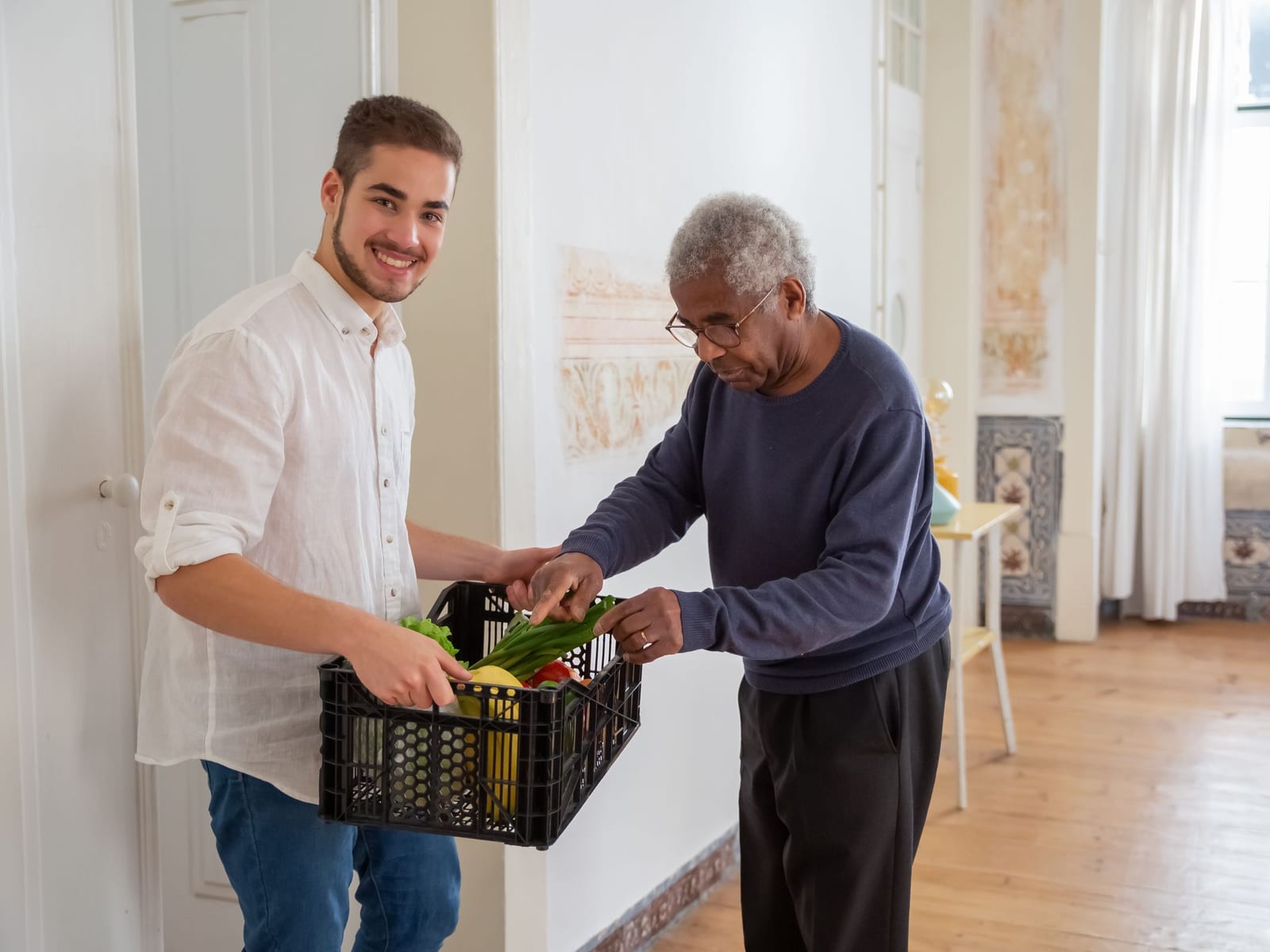A support professional and the man he supports unpack a crate of fresh groceries together in a bright hallway.