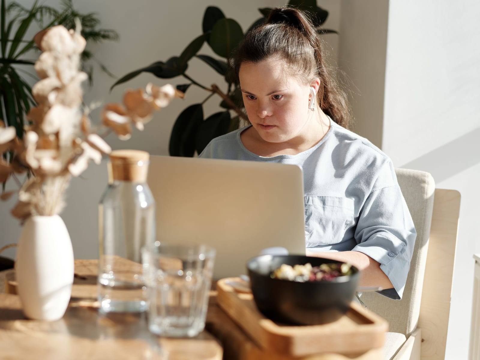 A young woman works at a laptop at her dining table, with plants and morning light around her.