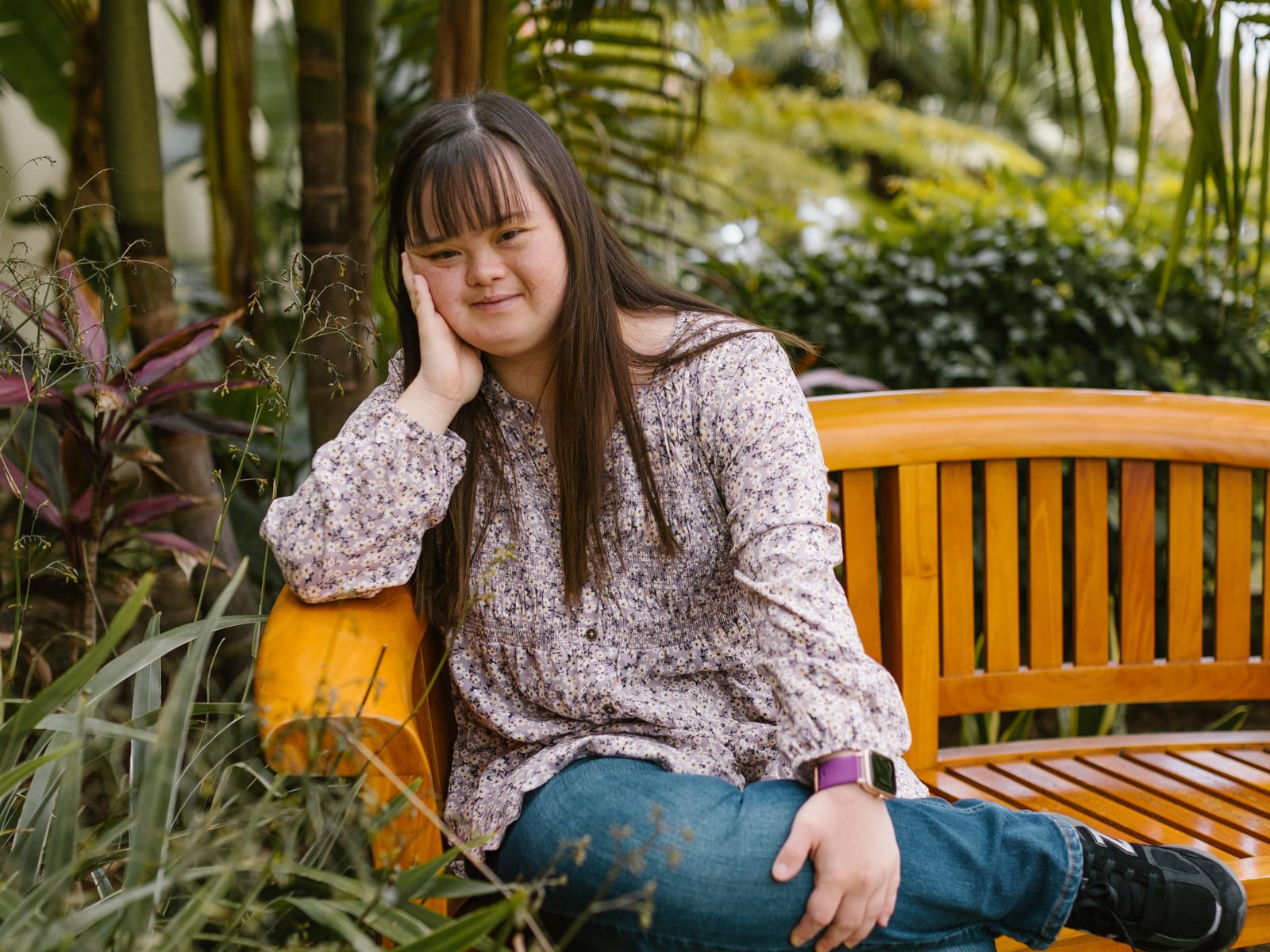A young woman rests on a wooden bench in a garden full of palms and flowering plants.