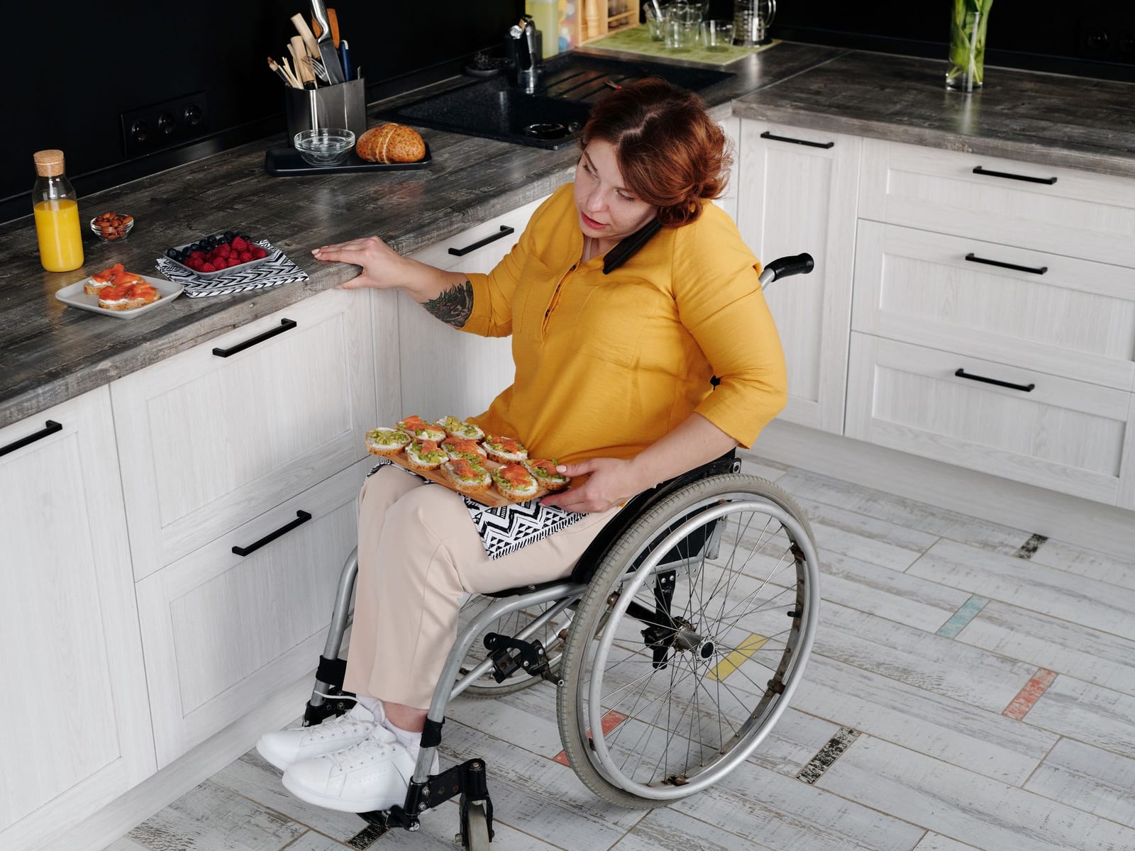 A woman in a wheelchair sets a tray of food she has just made on her lap in her own kitchen.